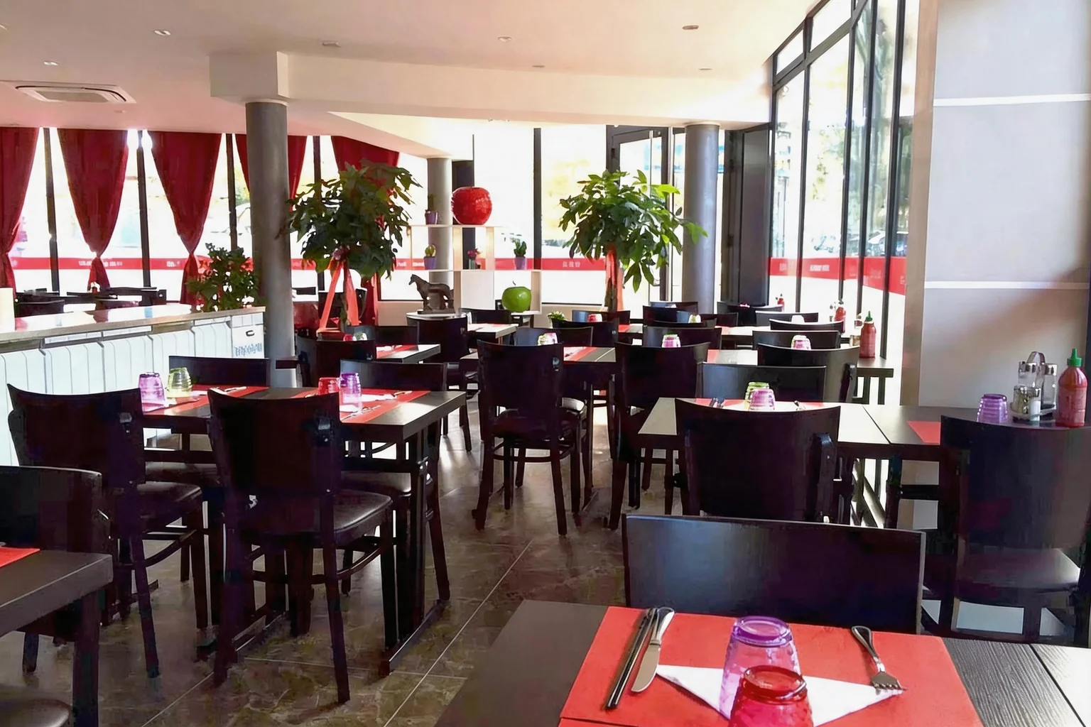 La salle : tables et chaises en bois sombre, sets de table rouges, rideaux rouges